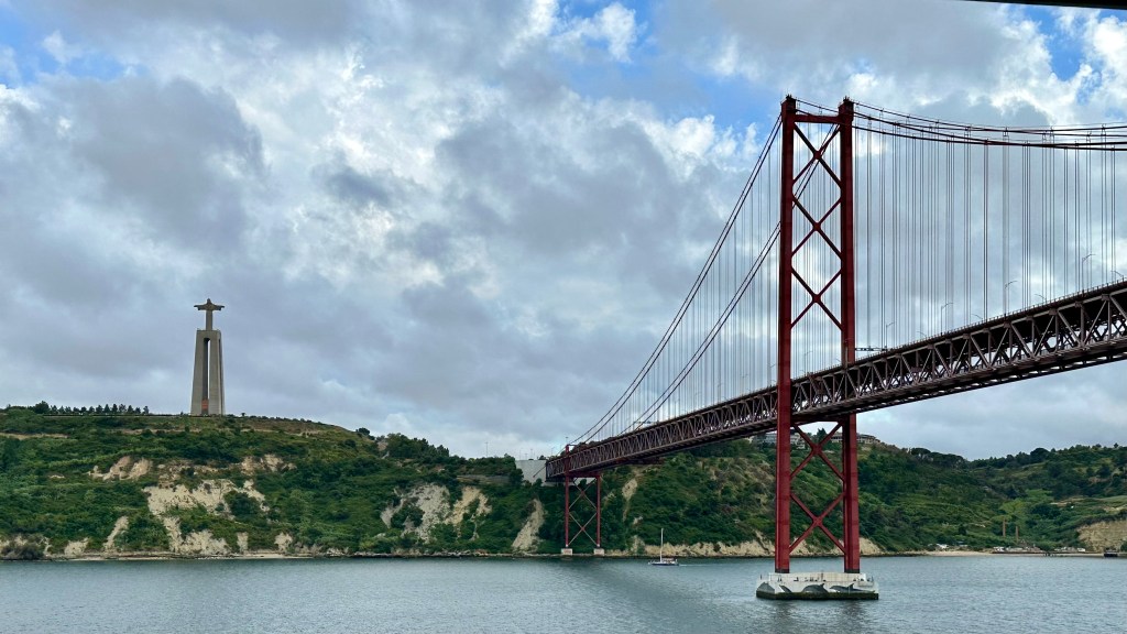 Taken from the private balcony of our cabin aboard the P&O Ventura as we slowly pulled away from Lisbon at the end of the day, this photograph captures one of the city's most iconic views. Dominating the right-hand side of the image is the striking red 25 de Abril suspension bridge, its elegant steel towers and countless suspension cables stretching gracefully across the River Tagus before disappearing beyond the edge of the frame. To the left, standing proudly on the opposite hillside, is the impressive Christ the King (Cristo Rei) monument, its outstretched arms overlooking Lisbon from its tall concrete pedestal. The green hills beneath the statue are broken by pale limestone cliffs that tumble down towards the calm water, while a small sailing yacht drifts quietly beneath the bridge, emphasising the immense scale of the engineering above. Overhead, a dramatic sky filled with thick grey clouds is broken by patches of brilliant blue, allowing shafts of soft evening light to illuminate parts of the landscape. The still water reflects the muted colours of the sky, creating a peaceful atmosphere as the Ventura begins its journey away from port. It was a wonderful moment to photograph from the comfort of our balcony cabin, offering a unique perspective that few viewpoints ashore could match.
