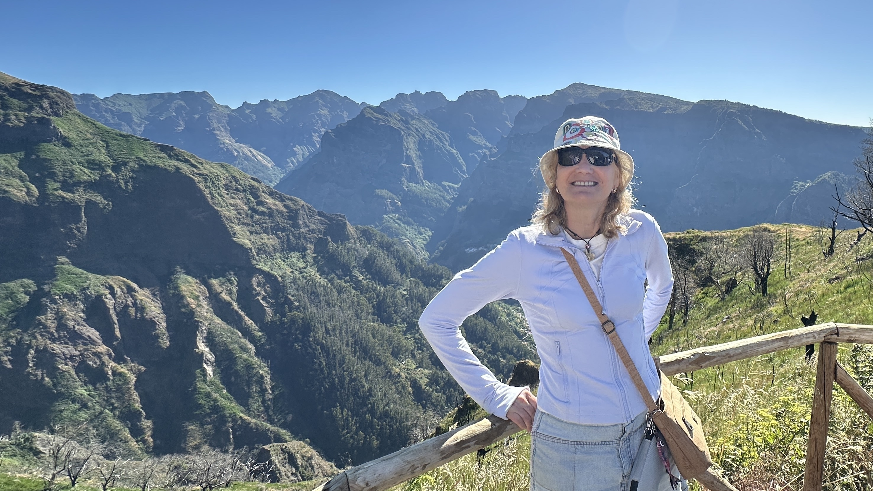 Subject (Sarah): Sarah is positioned slightly to the right of the center frame, leaning comfortably against a rustic, light-colored wooden safety log railing. She is smiling warmly at the camera, wearing dark sunglasses, a colorful bucket hat, a fitted light-lavender zip-up jacket, light denim bottoms, and a tan cross-body bag slung across her chest. Her right hand rests on her hip, framing her pose nicely. The bright, direct sunlight illuminates her face and jacket, making her stand out clearly against the background. Background & Landscape: Mountains: Behind Sarah is a breathtaking, expansive panorama of Madeira's dramatic mountain ranges under a completely clear, deep blue sky. Depth & Detail: On the left, steep, rugged green cliffs drop down into deep valleys lined with dense forests of dark green trees. In the far distance, a continuous ridge of jagged mountain peaks stretches across the horizon. Foreground: Surrounding the wooden railing where Sarah is standing are grassy patches and dry brush, with a few small dark tree branches or trunks visible along the right edge.